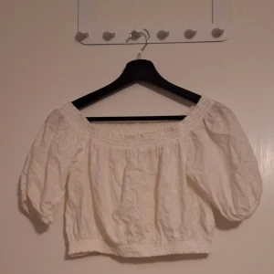 Vit broderad offshoulder blus - Superfin vit blus med broderade detaljer och små hålbroderier. Modellen är croppad med resår i midjan och vid axlarna för en offshoulder-look. Puffiga korta ärmar ger en romantisk vibe. Perfekt till jeans eller kjol för en söt och trendig stil.