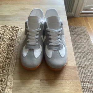 Gråvita sneakers med mockadetaljer - Snygga sneakers i vitt och ljusgrått med klassisk design. Ovandelen är i en mix av mocka och syntet, med vita snören och en brun gummisula som ger retrovibbar. Perfekta för dig som gillar stilrena och fräscha skor till vardags.