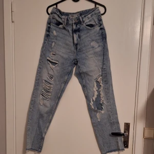 Ljusblå ripped jeans med hög midja - Säljer ett par ljusblå jeans med rejält slitna och trasiga detaljer framtill. Hög midja och raka ben, klassisk femficksmodell. Perfekta för dig som gillar en edgy och avslappnad stil. Materialet är klassisk denim i bomull.