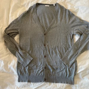 Grå kofta från Esprit med knappar - Snygg grå kofta från Esprit med v-ringad hals och bruna knappar framtill. Mjuk och tunn i bomullsmaterial, perfekt för lager-på-lager. Långa ärmar och ribbade muddar vid ärmslut och nederkant. Diskret logga broderad på bröstet.