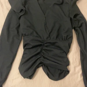 Svart oanvänd topp från Vero Moda - Svart oanvänd omlottopp från Vero Moda med lång ärm och snygg rynkad detalj framtill. Toppen har en v-ringad hals och är gjord i ett strukturerat, mjukt polyestertyg som ger en cool känsla. Perfekt att styla med jeans eller kjol för en trendig look.