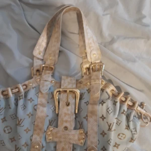 Louis Vuitton beige handväska med bälte - Säljer en Louis Vuitton handväska i beige och vit ish guld med klassiskt LV-mönster. Väskan har snygga detaljer i skinn med reptilmönster, guldfärgade spännen och bälten. Insidan är brun och har en präglad Louis Vuitton-logga. Perfekt för dig som vill sticka ut. Pris kan diskuteras! Skriv om ni undrar något😃. Nypris är runt 10-13 k men säljer för 5500 då den har vissa repor på handtag, och är lite skitig under och inuti men borde gå bort!