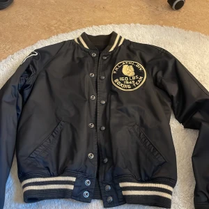 Svart bomberjacka Polo Ralph Lauren - Svart bomberjacka från Polo Ralph Lauren med broderade patches och text, bland annat 'New York Boxing Club' på ryggen och 'Boxing Team' på bröstet. Jackan har ribbade muddar med vita ränder, tryckknappar och coola detaljer som siffra på ärmen. Fickor både in och utsida.