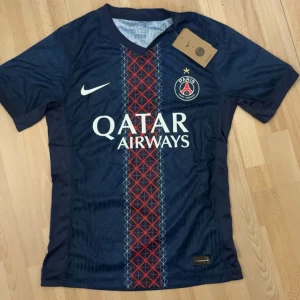 PSG matchtröja Nike marinblå - Paris Saint-Germain fotbollströja från Nike i marinblått med röd och vit grafisk rand mitt på bröstet. PSG-logga med stjärna, Qatar Airways tryck och vit Nike-swoosh. V-ringad hals och korta ärmar. Tillverkad i lätt, ventilerande material.