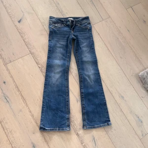 Blå bootcut jeans från Gina Tricot - Snygga blå jeans från Gina Tricot i storlek 152. Modellen har bootcut-ben, klassiska fem fickor och coola detaljer på bakfickorna med knappar. Jeansen har en normal passform och är tillverkade i slitstarkt denimtyg.