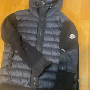 Svart Moncler cardigan med stickade ärmar - Storlek M Snygg svart dunjacka från Moncler med quiltad kropp och stickade ärmar. Jackan har huva, dragkedja framtill och praktiska fickor med dragkedja på ärmen. Perfekt för dig som vill ha en sportig och trendig look.