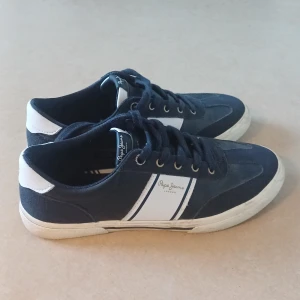 Mörkblå sneakers från Pepe Jeans - Snygga mörkblå och vita sneakers från Pepe Jeans med klassisk låg profil och platt sula. Skorna har snörning, rund tå och detaljer i både skinn och syntet. Perfekt för dig som gillar stilrena och sportiga vibes. Pepe Jeans-logga på sidan och tungan. OBS, köp via postnord.