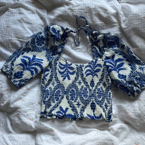 Blåvit blus med knyt från H&M XS - Supersöt blus från H&M i blått och vitt med tropiskt blommigt mönster. Toppen har puffiga trekvartsärmar, smockad struktur och knytning i nacken. Perfekt för dig som gillar unika detaljer och somriga vibes.