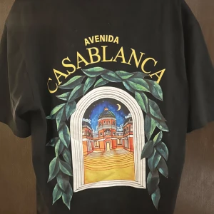 Svart t-shirt från Casablanca - Svart t-shirt från Casablanca med färgglad tryckt grafik på både bröstet och ryggen. Motivet föreställer en byggnad i fönsterformad ram med gröna blad och texten 'Avenida Casablanca' i gult. Klassisk passform och rund hals. Perfekt för dig som gillar unika prints.