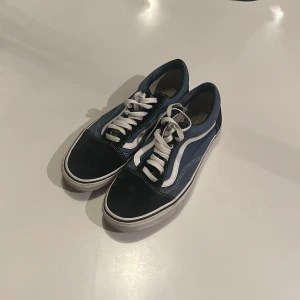 Blå och svarta Vans Old Skool sneakers - Klassiska Vans Old Skool sneakers i blått och svart med vit sula och vit sidosnöre. Skorna har rund tå, snörning och den ikoniska vågiga Vans-loggan på sidan. Ovandelen är i mocka och canvas, perfekt för en avslappnad streetstil. Pris kan självklart diskuteras!