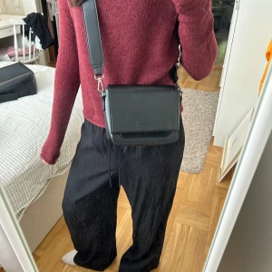 Svart axelväska från Carin Wester - Säljer en stilren svart axelväska från Carin Wester i slätt skinn med bred justerbar axelrem. Väskan har magnetlås, innerfack och en snygg detalj i mocka på framsidan. Liten men rymlig väska!!