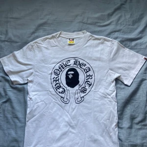Vit BAPE x Chrome Hearts t-shirt M - Säljer en vit t-shirt från BAPE i samarbete med Chrome Hearts. Stor svart print med ikoniska BAPE-apan och Chrome Hearts-logga fram, samt Baby Milo och korsdetalj bak. Klassisk rund hals och korta ärmar. Materialet är mjuk bomull, perfekt för streetwear.Ikke autentiskt