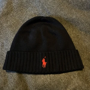 Mörkblå stickad mössa från Polo Ralph Lauren - nyskick!  - Snygg svart stickad mössa från Polo Ralph Lauren med klassisk röd broderad logga framtill. Mössan har uppvikt kant med ribbad struktur som ger en schysst look. Perfekt för kalla dagar och enkel att matcha med din stil.