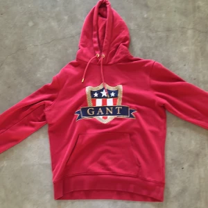 Röd hoodie från GANT med broderad logga - Säljer en röd hoodie från GANT med stor broderad logga och sköld på bröstet. Tröjan har huva med snörning, guldfärgade detaljer och en stor magficka. Mjuk bomullskvalitet och långärmad passform, perfekt för chill dagar. Den har ett litet hål på känguru fickan som man ser på bild nr3 men går att laga. 🫣köpt för 1200kr🫣 🍀Säljer för 630kr pga av det lilla hålet🍀.    Har en till fast grå som ni kan se på min andra annons! Om ni köper båda tillsamans får ni båda för 1100kr!🍀🍀