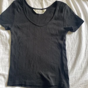 Svart ribbad topp från H&M - En svart ribbad topp från H&M med korta ärmar och rund halsringning. Toppen har diskreta vertikala ränder i strukturen som ger en snygg detalj. Perfekt att styla med jeans eller kjol för en enkel och clean look.