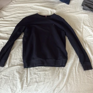 Mörkblå sweatshirt från Tommy Hilfiger - En stilren mörkblå sweatshirt från Tommy Hilfiger med diskret logga på bröstet. Tröjan har rund hals, långa ärmar och ribbade muddar. Perfekt för en clean och klassisk look.
