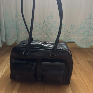 Svart axelväska i skinn med fickor - Snygg svart axelväska i glansigt skinn med två stora fickor framtill och långa axelband. Väskan har dragkedja upptill och flera innerfack, perfekt för att organisera mobilen och småsaker. Stilren och praktisk design som passar till många outfits. Får plats med 1 liter vattenflaska annars kläder.