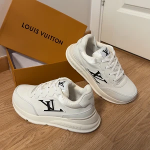 Louis Vuitton skor - Snygga vita sneakers från Louis Vuitton med tydlig svart LV-logga på sidan och tungan. Skorna har rund tå, platt sula och är tillverkade i skinn och syntet. Klassisk snörning och diskreta detaljer gör dem till ett stilrent val för dig som gillar exklusiva märken.