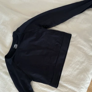 Svart croppad träningströja - Svart croppad långärmad träningströja från H&M med diskret prickigt mönster och tumhål i ärmsluten. Tröjan har ribbade muddar och är gjord i ett stretchigt syntetmaterial som andas. Perfekt för gymmet eller löprundan.