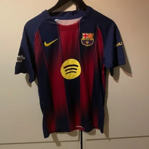 FC Barcelona Lamine Yamal #10 matchtröja - Säljer en FC Barcelona matchtröja med Lamine Yamal och nummer 10 på ryggen. Tröjan är kortärmad med klassiska blå och röda vertikala ränder, gula detaljer och klubbmärke på bröstet. Tillverkad i lätt och ventilerande material, perfekt för fotboll eller häng med polarna.