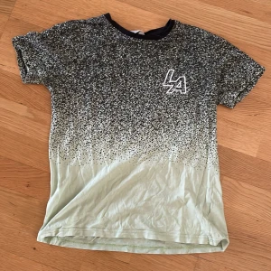 Svart och ljusgrön t-shirt från H&M - Cool t-shirt från H&M med svart och ljusgrön färg som tonar ut i varandra. Prickigt mönster över hela tröjan och en vit LA-logga på bröstet. Rund halsringning och korta ärmar. Perfekt för dig som gillar streetstyle och grafiska prints.