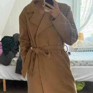 Camel kappa med bälte och fickor - Snygg camelbrun kappa med bälte i midjan och stora slag. Lång modell med fickor på sidorna och klassisk krage. Perfekt för dig som vill ha en stilren och tidlös look under höst och vinter. Materialet känns mjukt och värmande.