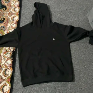 Svart hoodie från Polo Ralph Lauren - Svart hoodie från Polo Ralph Lauren med klassisk huva och lång ärm. Tröjan har en liten vit Polo-logga broderad på bröstet och en stor magficka framtill. Perfekt för chill dagar och enkel att matcha med jeans eller joggers.