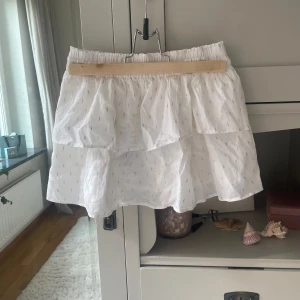 Vit volangkjol från Gina Tricot - Supersöt vit kortkjol från Gina Tricot med volanger och små guldiga detaljer. Kjolen har resår i midjan för skön passform och är gjord i mjukt bomullstyg. Perfekt för dig som gillar en ljus och glittrig look med extra volym.