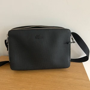 Svart axelväska från Lacoste - Snygg svart axelväska från Lacoste med diskret krokodillogga framtill. Väskan har justerbar axelrem, silverfärgad dragkedja och grönt foder med innerfack. Tillverkad i tålig syntet med strukturerad yta. Perfekt för att bära det viktigaste med stil.