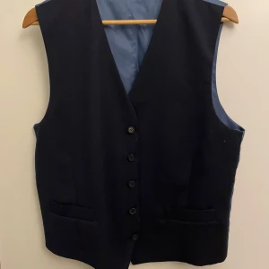 Mörkblå kostymväst från Dressman - Snygg mörkblå kostymväst från Dressman med klassisk v-ringning och fem knappar framtill. Västen har två fickor och är fodrad med blått tyg på insidan. Perfekt för att lyfta din stil till nästa nivå.