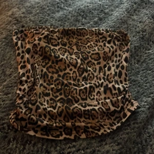 Leopardmönstrad bandeau tubtopp - Säljer en leopardmönstrad bandeau tubtopp i mjukt och glansigt syntetmaterial. Toppen har en beige och brun bas med svarta leopardfläckar och är axelbandslös, perfekt för att ge din outfit en edgy vibe.