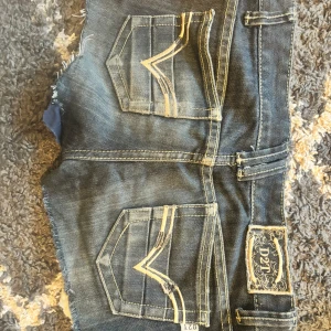Blå jeansshorts från D2T - Säljer ett par blå jeansshorts från D2T med råa, fransiga kanter och snygga kontrastsömmar. Shortsen har klassiska fem fickor och coola detaljer på bakfickorna. Passar dig som gillar streetstyle och denim med attityd.