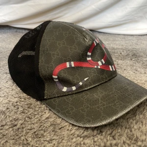 Gucci keps med orm och GG-mönster - Snygg keps från Gucci med klassiskt GG-mönster i mörkgrått och svart. Framsidan pryds av en röd, vit och svart randig orm. Baksidan har svart mesh och justerbart band med röd och marinblå rand. Perfekt statement-piece för dig som vill sticka ut.