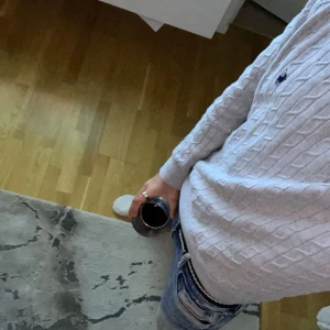 Ljusgrå kabelstickad tröja Polo Ralph Lauren - Säljer en ljusgrå kabelstickad tröja från Polo Ralph Lauren med klassisk blå logga på bröstet. Tröjan har rund hals, lång ärm och ribbade muddar. Perfekt för dig som gillar stilren och tidlös design med en preppy touch. Den har ett litet oljig fläck som går bort enkelt med diskmedel eller potatis mjöl 