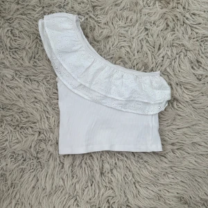 Vit one shoulder topp med volang - Superfin vit one shoulder topp med bred broderad volang och smal axelrem. Toppen är croppad och har en söt spetsdetalj på volangen. Perfekt för sommarens varma dagar och ger en fräsch look till jeans eller kjol.