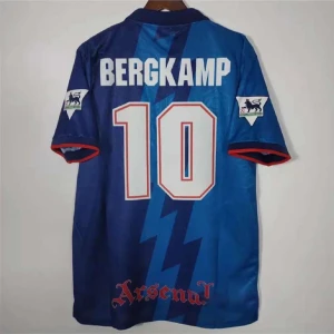 Arsenal Bergkamp 10 blå fotbollströja - Retro Arsenal fotbollströja med Bergkamp 10 på ryggen. Tröjan är blå med mörkare blå blixtmönster, röda detaljer vid ärmslut och klubbnamnet tryckt längst ner. Premier League-logga på ärmarna och klassisk krage. Perfekt för fans av Bergkamp och Arsenal.