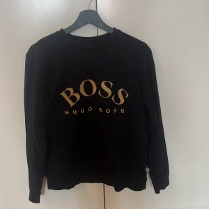 Svart sweatshirt från Hugo Boss - Svart sweatshirt från Hugo Boss med stort guldigt BOSS-tryck på bröstet. Klassisk rund halsringning och långa ärmar. Mjuk och skön bomullskänsla, perfekt för en stilren och avslappnad look.