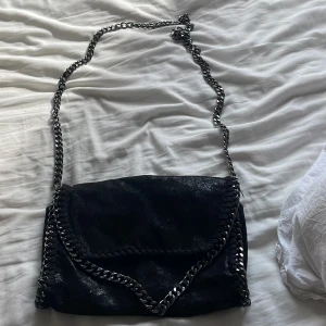 Svart axelväska med kedja - Säljer en svart axelväska med glansig finish och grov silverfärgad kedja som axelrem. Väskan har dekorativa kedjedetaljer längs kanterna och magnetlås. Insidan är fodrad med leopardmönster. Perfekt för dig som gillar edgy och trendig stil.