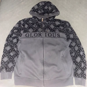 Grå hoodie Glorious med mönster - Snygg grå hoodie från Glorious med svart grafiskt mönster på ärmar och huva. Dragkedja framtill, broderad logga över bröstet och justerbar huva med snören. Perfekt för dig som gillar streetwear vibes.