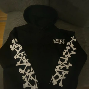 Svart Amiri hoodie med benmönster - Svart hoodie från Amiri med vita benmönster längs båda ärmarna och tryckt logga på bröstet. Tröjan har huva och är tillverkad i 100% bomull. Perfekt för dig som gillar streetwear med edge.