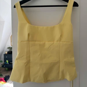 Gul linnetopp med rosett  - Supersöt ljusgul linnetopp med breda axelband, fyrkantig ringning och en stor rosett i ryggen. Toppen har en snygg cut out-detalj baktill och är lätt utsvängd nertill. Perfekt för soliga dagar och ger en fräsch vibe till din outfit.