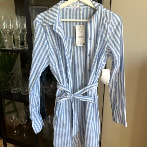 Randig skjortklänning från NA-KD - Snygg skjortklänning från NA-KD i blåvitt randigt mönster. Klänningen har långa ärmar, knappar framtill och ett knytbälte i midjan. Tillverkad i en skön linnemix som passar perfekt för varmare dagar. Klassisk krage och avslappnad passform.