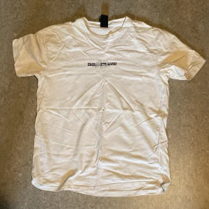 Vit t-shirt med draktryck från Black Squad - Vit t-shirt från Black Squad med avslappnad passform. Framsidan har en liten svart text, medan baksidan har ett stort, detaljerat draktryck och texten 'paranoid'. Tillverkad i mjuk bomull, perfekt för dig som gillar streetstyle och coola prints.