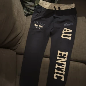 Mörkblå mjukisbyxor med tryck - Mörkblå mjukisbyxor med bred, ljusgrå ribbad midja och snörning. Snyggt tryck med texten 'Denim Authentic' längs benet och 'Athletic New York' vid fickan. Perfekta för chill eller träning, med mjukt material och avslappnad passform.