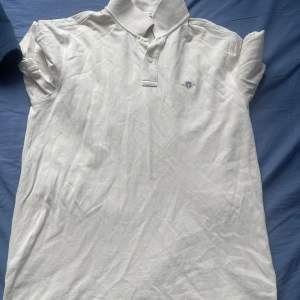 Vit pikétröja från GANT i bomull - Klassisk vit pikétröja från GANT med broderad logga på bröstet. Tröjan har korta ärmar, krage och knappar framtill. Tillverkad i mjuk bomull som känns skön mot huden. Perfekt för en clean och stilren look.