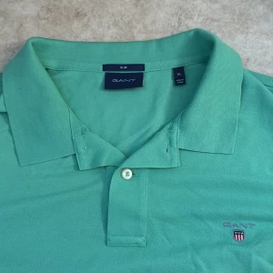 Grön pikétröja från GANT, slim fit XL - Snygg grön pikétröja från GANT i slim fit-modell. Klassisk krage, kort ärm och två knappar framtill. Broderad GANT-logga med sköld på bröstet. Tillverkad i mjuk bomull som andas, perfekt för vår, höst och sommar.
