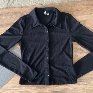 Svart ribbad skjorta med knappar - Stilren svart skjorta med ribbad struktur och klassisk krage. Skjortan har långa ärmar och knäppning framtill med svarta knappar. 