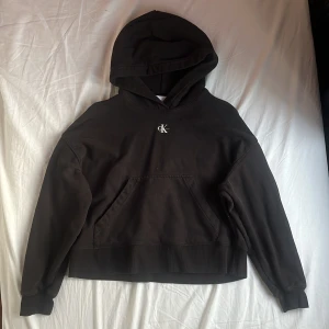Svart hoodie från Calvin Klein Jeans - Svart hoodie från Calvin Klein Jeans med broderad CK-logga på bröstet. Klassisk känguruficka framtill, huva och ribbade muddar. Skön passform och tillverkad i mjuk bomull. Perfekt för en chill och stilren look. Nypris 1195 kr.