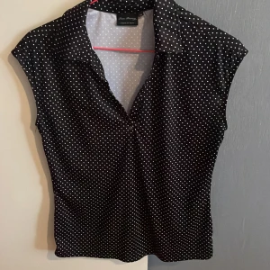 Svart prickig blus från Func Factory - Snygg svart blus med vita prickar från Func Factory. Modellen har krage, korta ärmar och en figurnära passform. Perfekt för dig som gillar retrovibbar och vill sticka ut med ett klassiskt mönster.