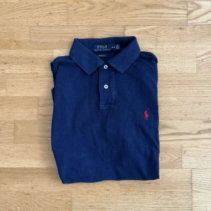 Ralph Lauren piké  - Klassisk marinblå pikétröja från Ralph Lauren med röd broderad logga på bröstet.  Tillverkad i mjuk bomull som är skön mot huden och passar perfekt till en avslappnad stil. 